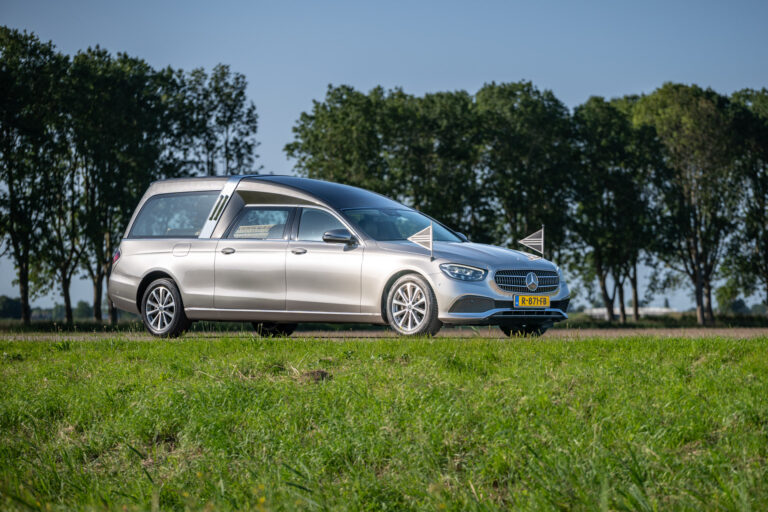 Zilvergrijze rouwauto met rouwvlaggetjes langs een bomenrij in de polder